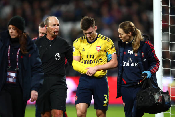 LONDON, ENGLAND - DECEMBER 09: Referee Mike Dean helps Kieran Tierney of Arsenal leave the pitch following an injury during the Premier League match between West Ham United and Arsenal FC at London Stadium on December 09, 2019 in London, United Kingdom. (Photo by Julian Finney/Getty Images)
