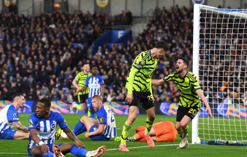 Arsenal star hits 14 goals/assists in last 13 games 3 Arsenal players in fluorescent yellow and black kits are celebrating a goal, with Kai Havertz seen cheering and being congratulated by teammate Jorginho after scoring. Behind them, Brighton players in blue and white stripes sit or kneel on the pitch, displaying expressions of disappointment and frustration. The goalkeeper lies on the ground in an orange kit, having been unable to stop the goal. The stadium is filled with spectators in the background, capturing a moment of contrasting emotions between the two teams.