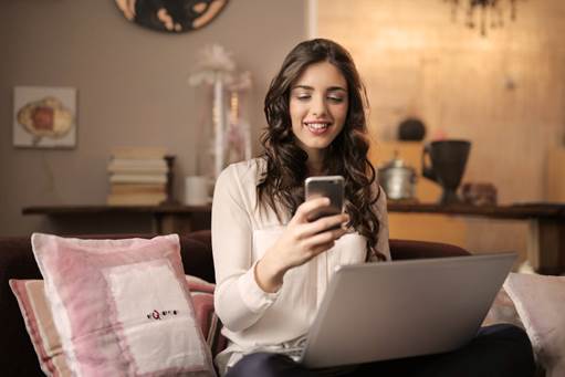 woman looks at her phone while she sits on a sofa in front of a laptop generic