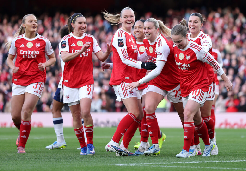 Arsenal Women players in red and white home shirts celebrating together on the pitch, with a packed Emirates Stadium crowd in the background.