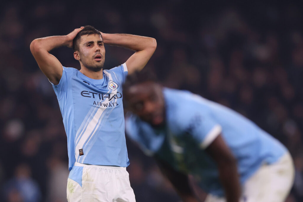Rodri reacts with both hands on his head during a Manchester City match, with a blurred teammate bent over in the background.