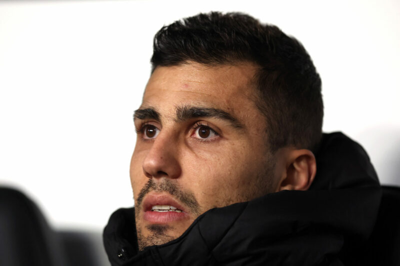 Close-up of Rodri in a black padded coat, looking on from the sidelines with a serious expression.