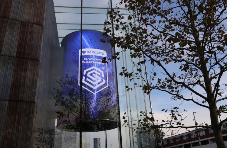 Large curved digital screen on the exterior of a modern glass building displaying the Barclays FA Women’s Super League logo, with bare tree branche...