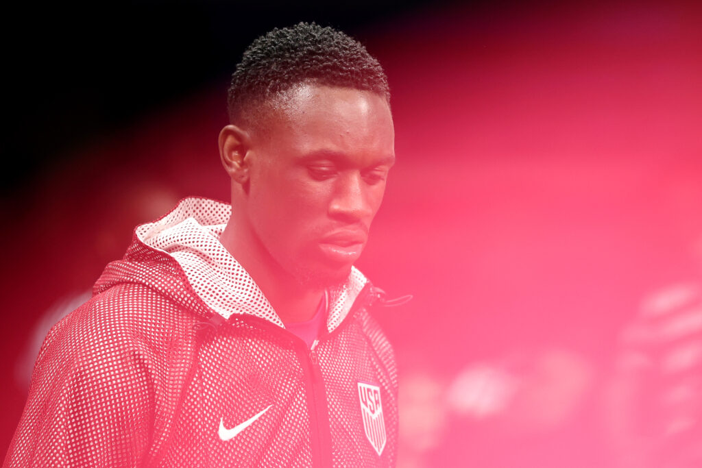Close‑up of United States forward Folarin Balogun in a red and white patterned USA warm‑up jacket, partially obscured by a soft pink blur in the foreground