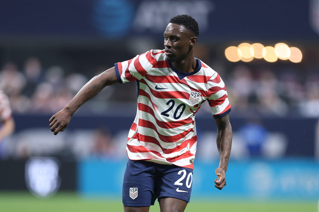United States forward Folarin Balogun, wearing the number 20 shirt in a red‑and‑white striped USA home kit, runs during a match with the stadium crowd blurred in the background.