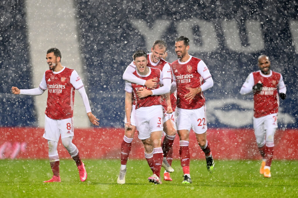 Kieran Tierney celebrates scoring for Arsenal as he's hugged by Rob Holding. It is snowing, he is in short sleeves, while all his teammates are in long sleeves