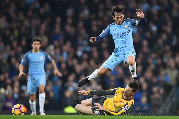 Arsenal's French defender Laurent Koscielny tackles Manchester City's Spanish midfielder David Silva (up) during the English Premier League football match between Manchester City and Arsenal at the Etihad Stadium in Manchester, north west England, on December 18, 2016. / AFP / Paul ELLIS / RESTRICTED TO EDITORIAL USE. No use with unauthorized audio, video, data, fixture lists, club/league logos or 'live' services. Online in-match use limited to 75 images, no video emulation. No use in betting, games or single club/league/player publications. / (Photo credit should read PAUL ELLIS/AFP/Getty Images)