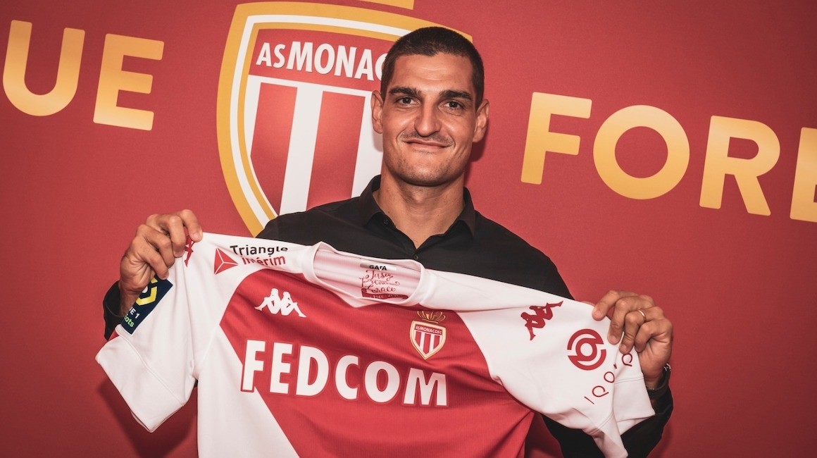 a picture of Vito Mannone holding an AS Monaco shirt in front of the AS Monaco badge on the wall behind him