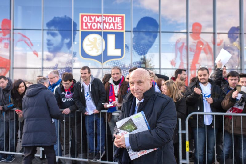 French former player Fleury Di Nallo stands in front of the Parc de l'Olympique Lyonnais on February 14, 2016 in Decines-Charpieu, central eastern France, as he inaugurates with OL's president "The avenue of lights" before the French L1 football match between Olympique Lyonnais (OL) and Stade Malherbe Caen (SMC). Fleury Di Nallo is seen wearing a suit and holding a folder, surrounded by people. (Photo credit ROMAIN LAFABREGUE/AFP via Getty Images)