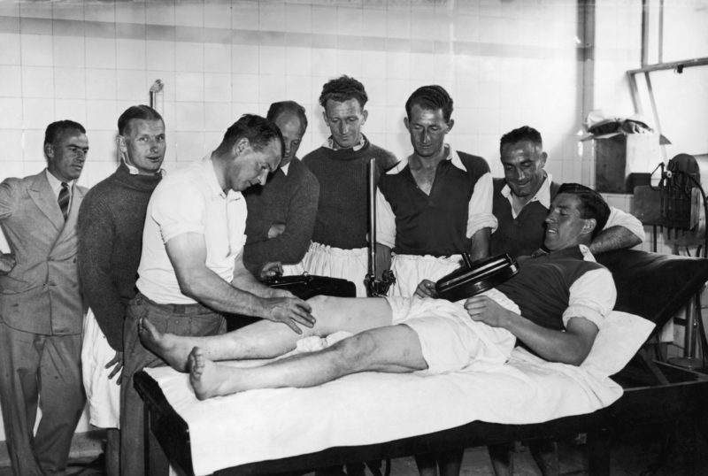 Arsenal trainer Tom Whittaker treating Alfred Kirchen with a short wave diathermy machine in the changing room at the Arsenal Stadium, Highbury, while his team mates look on, 30th August 1937. From lef to right Cliff Bastin, George Male, Herbert Roberts, John Crayston and Wilf Copping. (Photo by Topical Press Agency/Hulton Archive/Getty Images)