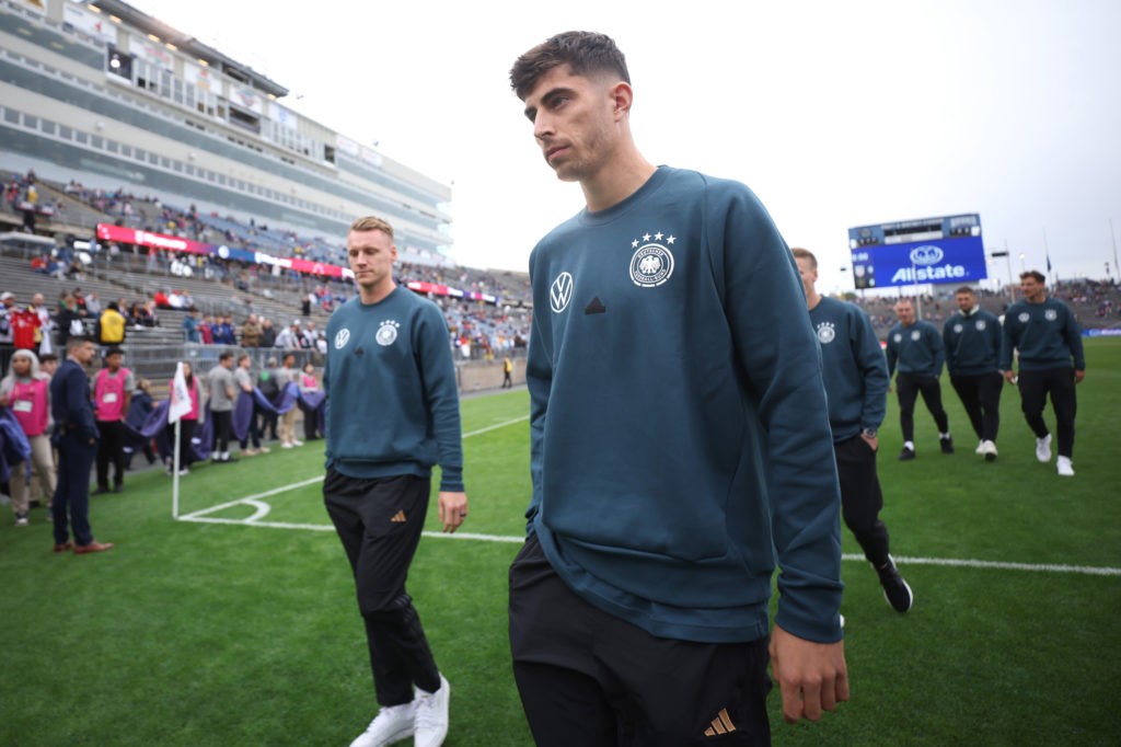 Kai Havertz wearing a Germany training jumper, walking on a football pitch with teammates and a stadium backdrop.